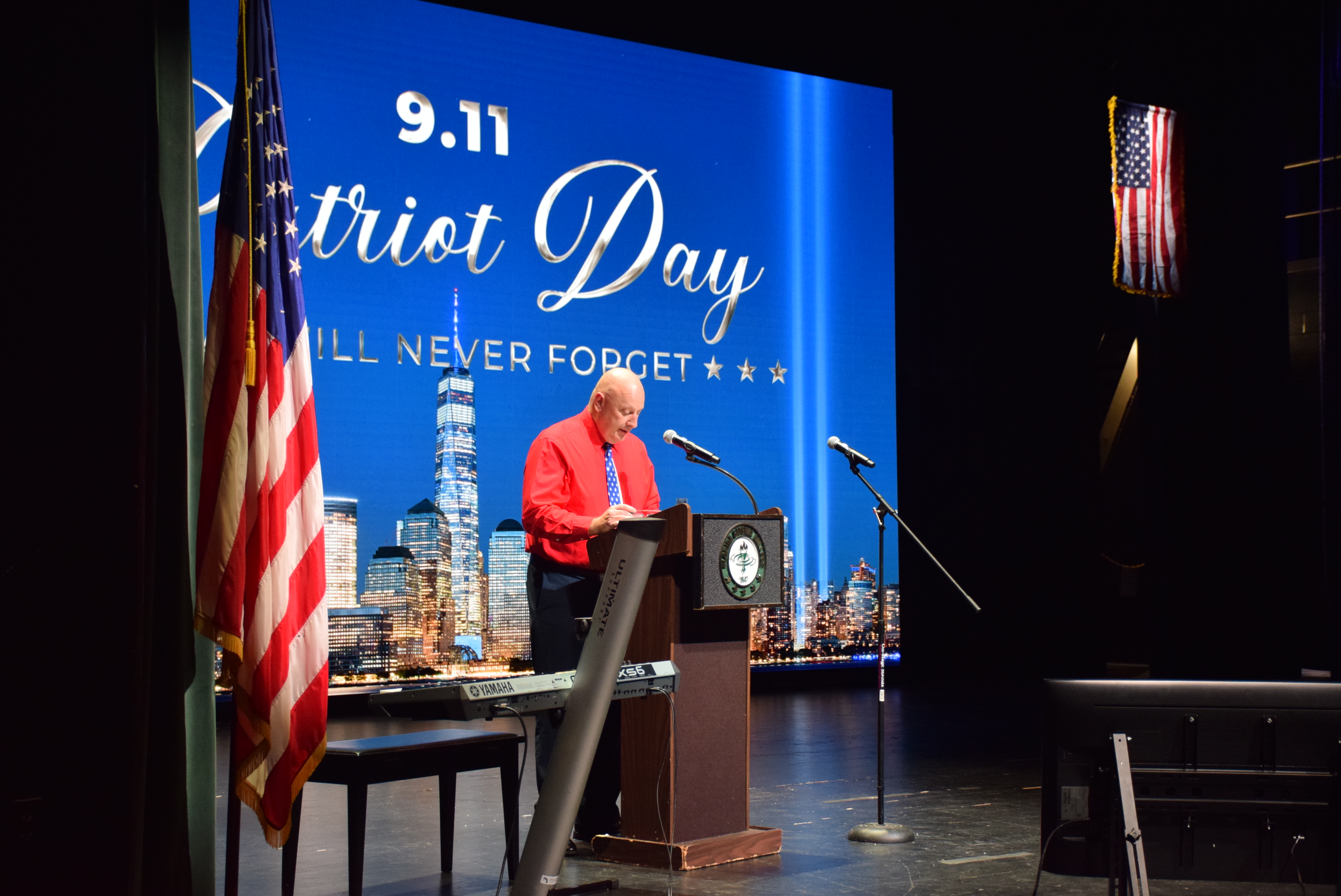 A speaker at a podium during a 9/11 Patriot Day memorial ceremony. An American flag stands nearby, and a projection screen behind displays 