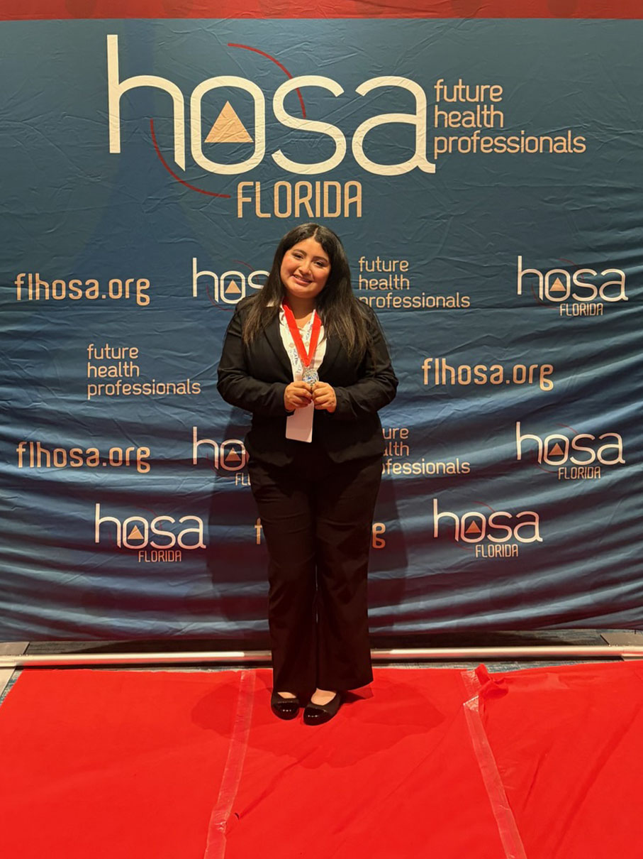 Portrait of a woman standing on a red carpet in front of a HOSA Florida backdrop, smiling and holding a medal while dressed in professional attire.